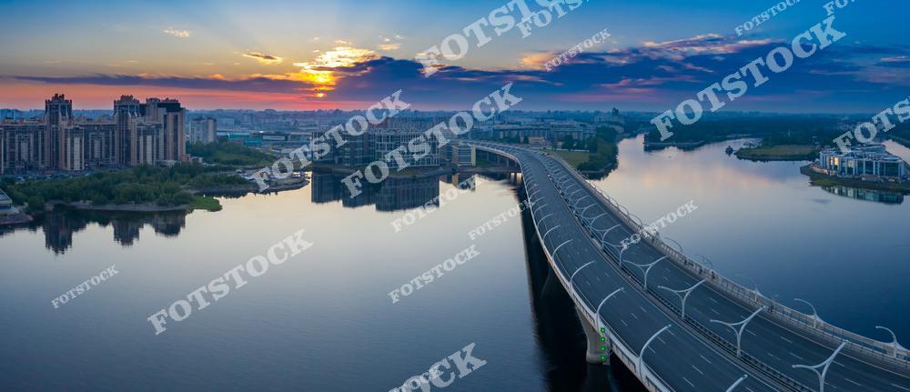 Фотография утренней панорамы Санкт-Петербург, Россия, снятая с высоты на рассвете. На изображении показан просыпающийся город: реки Санкт-Петербурга, мост через водную гладь и скоростная автомагистраль, проходящая по мосту. Фотоснимок передаёт мягкий свет восходящего солнца, отражения в воде и масштаб городской инфраструктуры. Фото подчёркивает характерный облик Петербурга как города рек и мостов, сочетание транспортной архитектуры и природного пространства. Иллюстрация может использоваться как изображение, картинка или слайд, посвящённые утренним видам и панорамам российских городов.